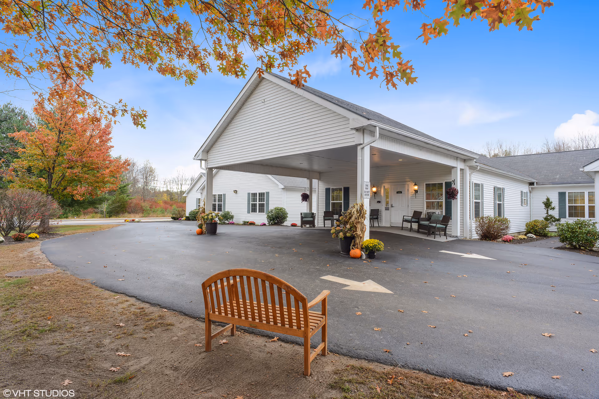 Exterior view of a single-story white building with a covered entrance and a driveway. There are autumn trees with orange leaves in the background, a wooden bench in the foreground, and some potted plants and pumpkins near the entrance.