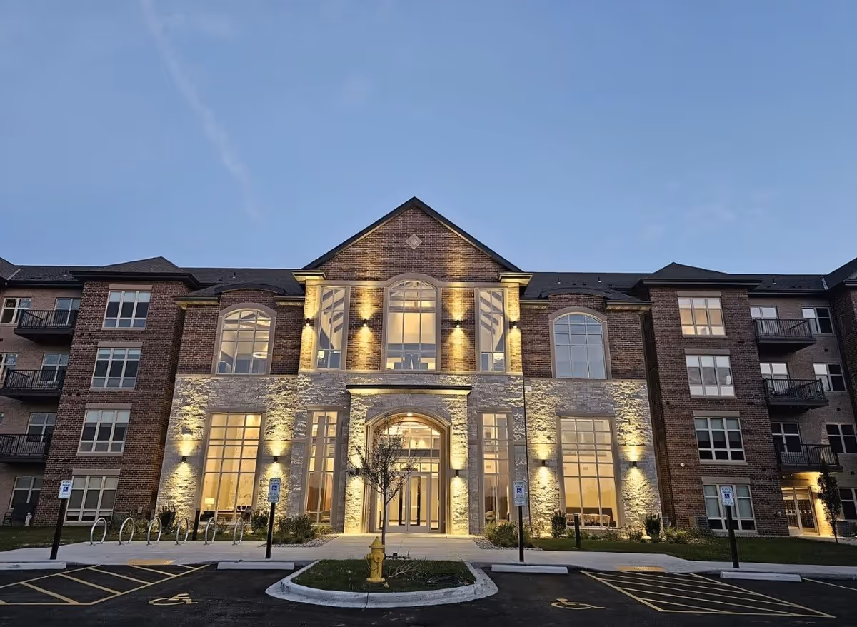 Front exterior view of a modern three-story brick and stone building with large windows and illuminated entrance at dusk. The building has balconies on the sides and accessible parking spaces in front.