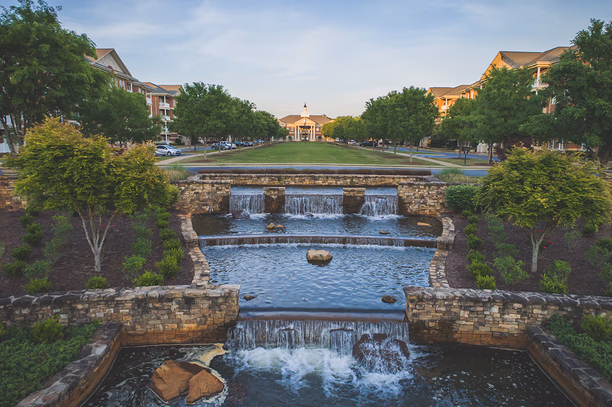 A landscaped outdoor area at Linville Court at The Cascades Verdae featuring a tiered stone water feature with cascading waterfalls, surrounded by greenery and trees, with residential buildings and a central building with a cupola in the background under a clear sky.