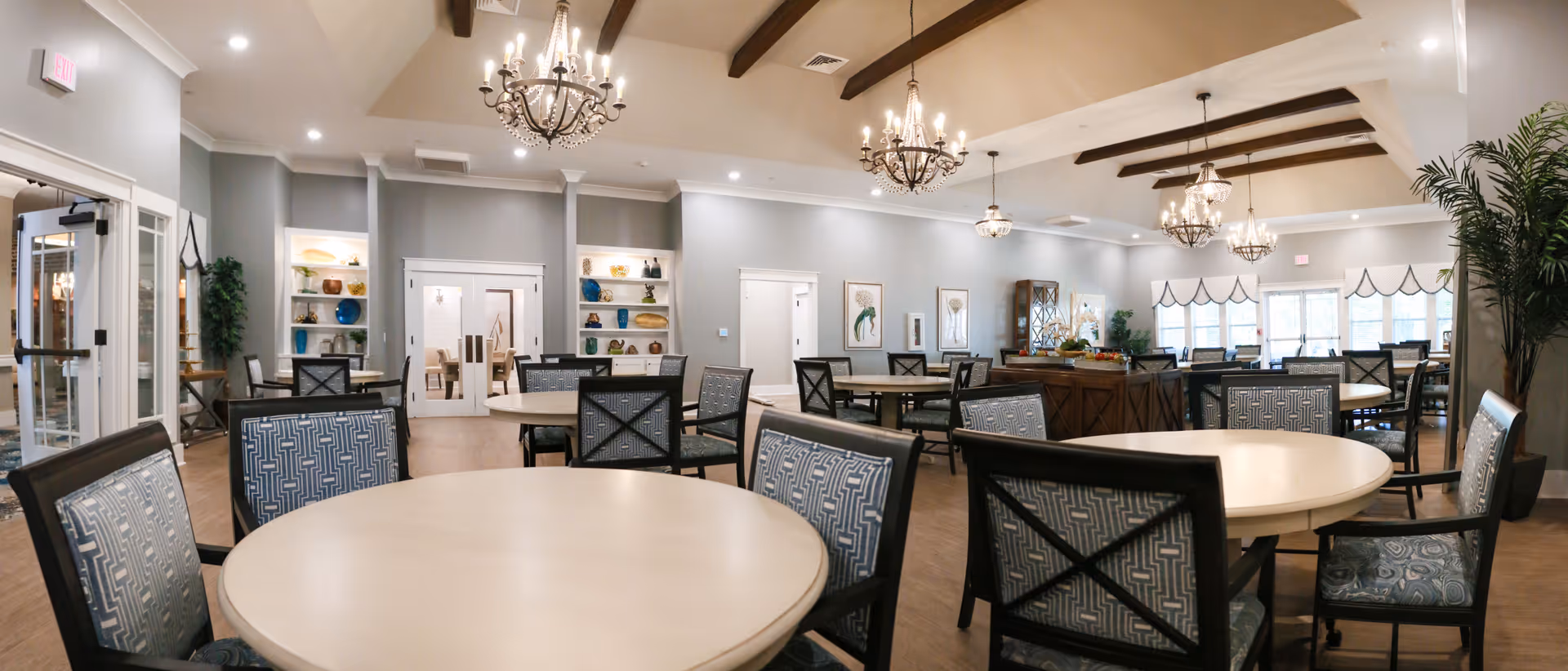 Bright senior living dining area with round tables, chairs, and decorative chandeliers.