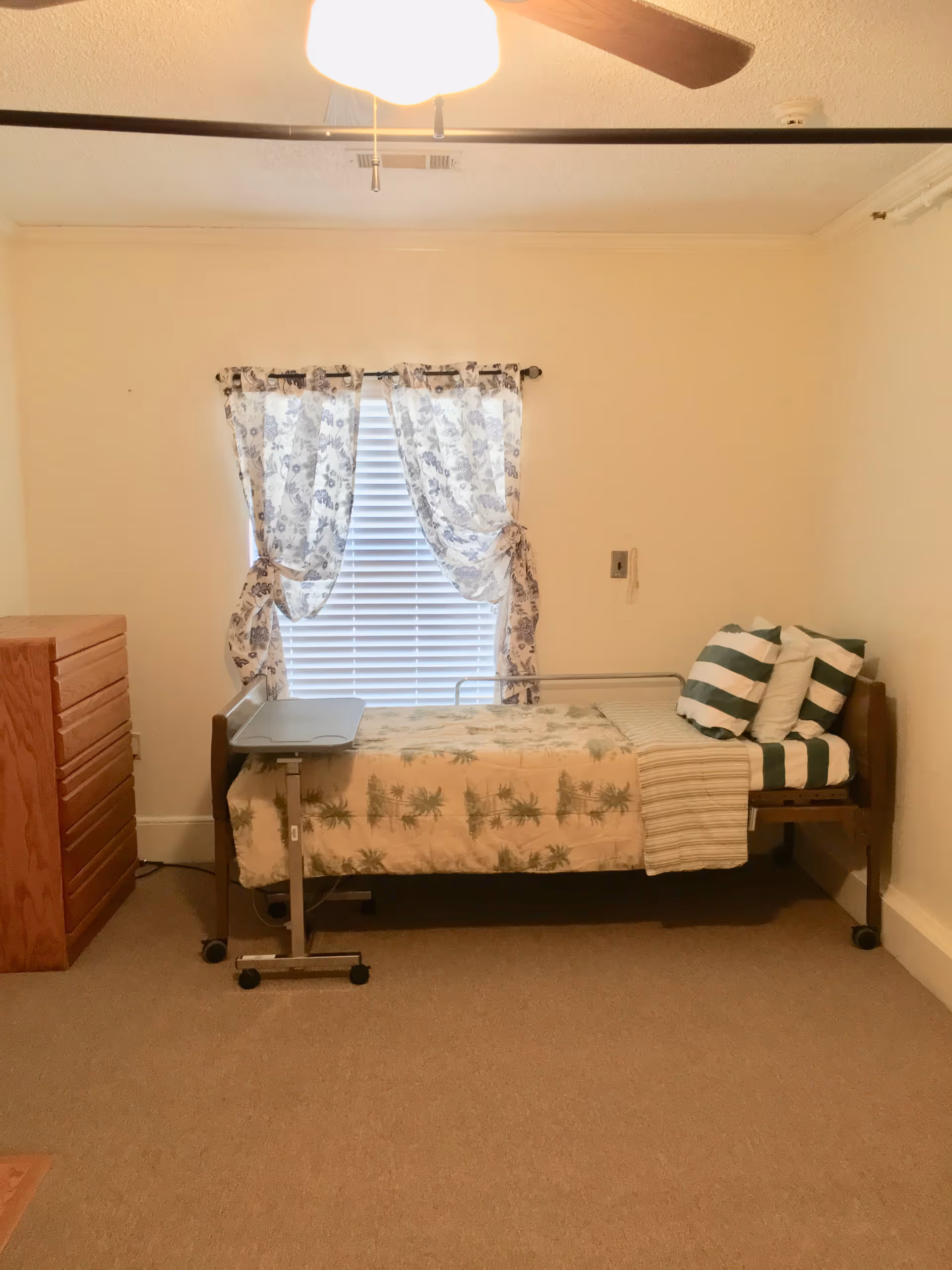 A single bed with palm-printed quilt, floral curtains, and a dresser in a small bedroom.