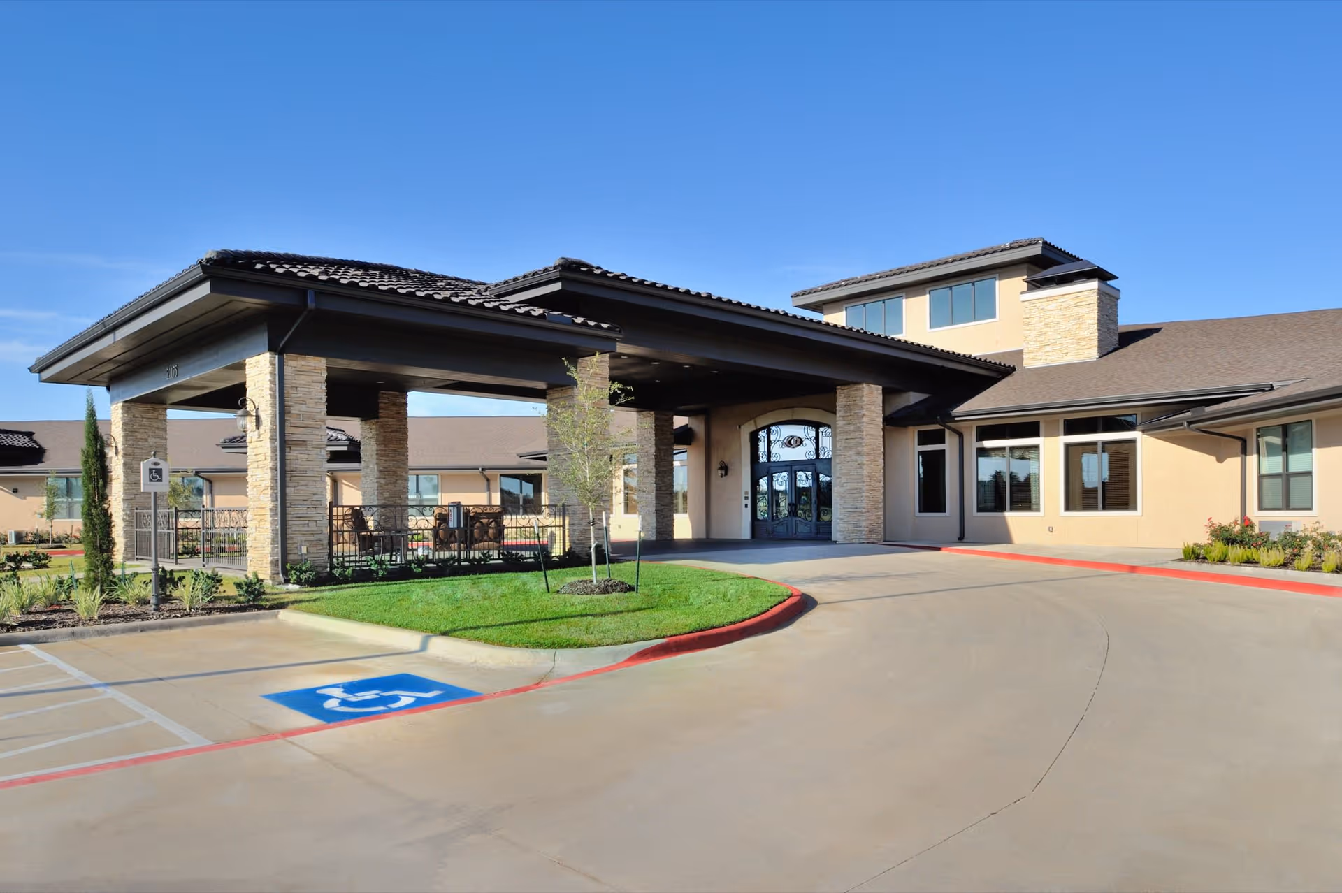 Exterior view of Clayton Oaks Living facility entrance with a covered drop-off area supported by stone pillars, a wheelchair accessible parking space in front, and a clear blue sky above.
