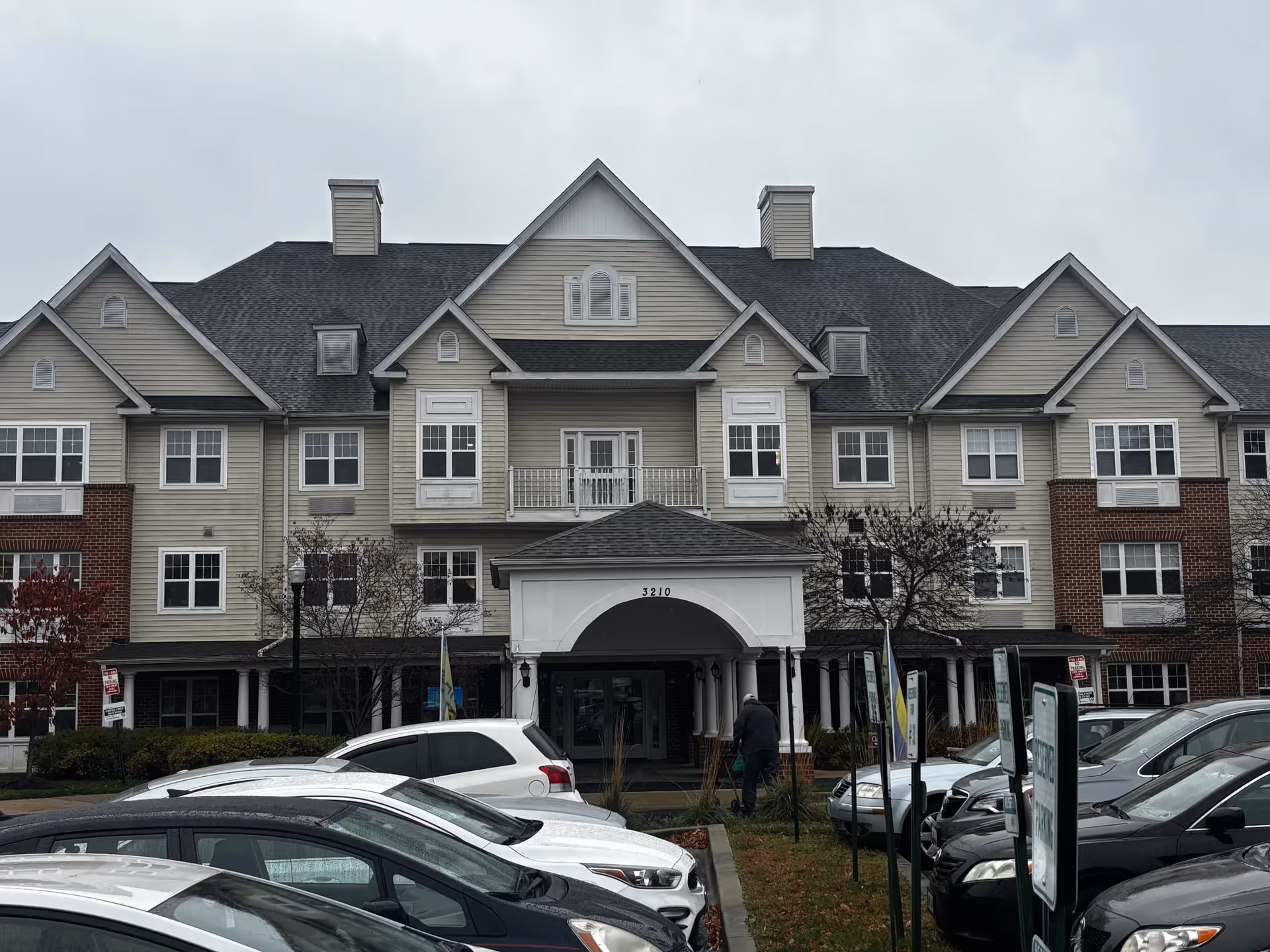 Front exterior of a multi-story senior living building with a covered entrance, balconies, and parked cars in the foreground.
