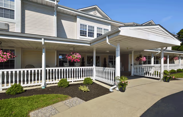 Covered entrance of a two-story senior living building with white railings, hanging pink flower baskets, and a paved walkway.