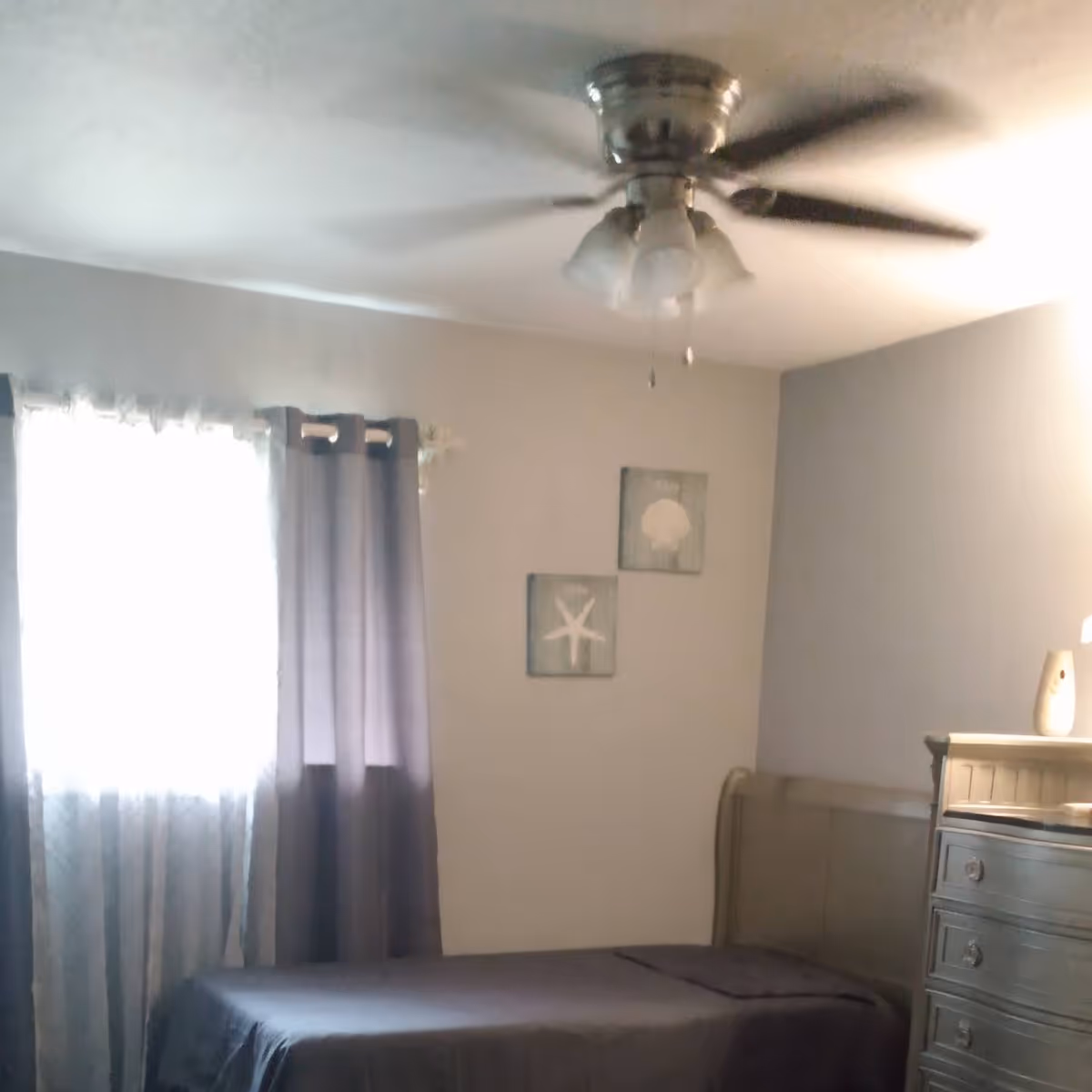 A tidy bedroom in a senior living facility with bed, dresser, window, curtains, and ceiling fan.