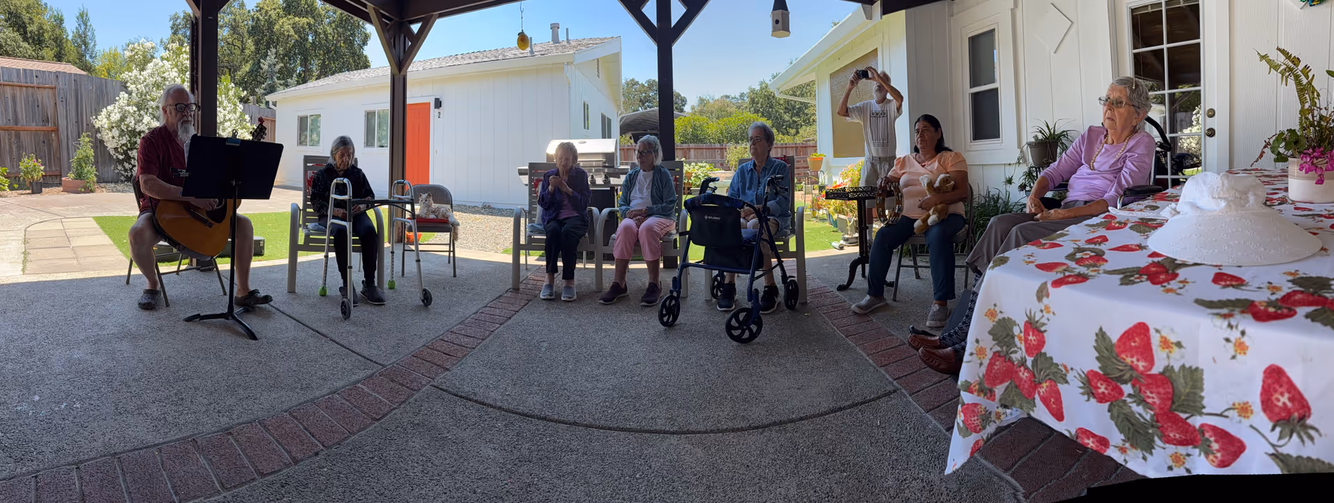Group of seniors gather outdoors under a covered patio, some in wheelchairs, listening to guitar.