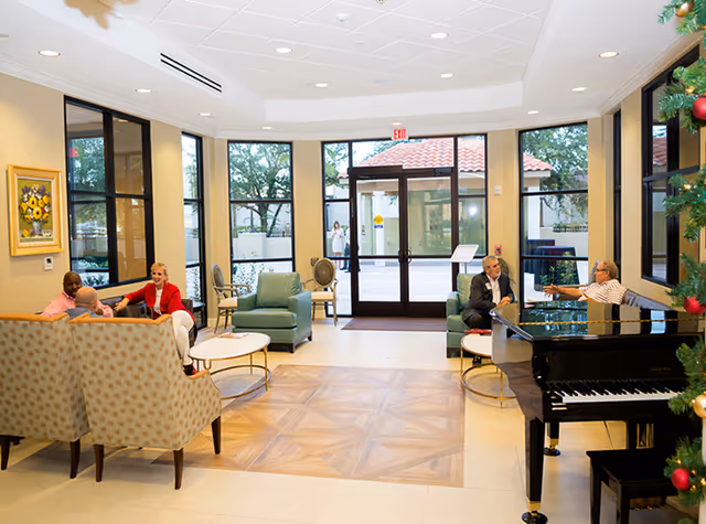 Senior residents relax in a bright lobby with seating, a piano, and festive decor.