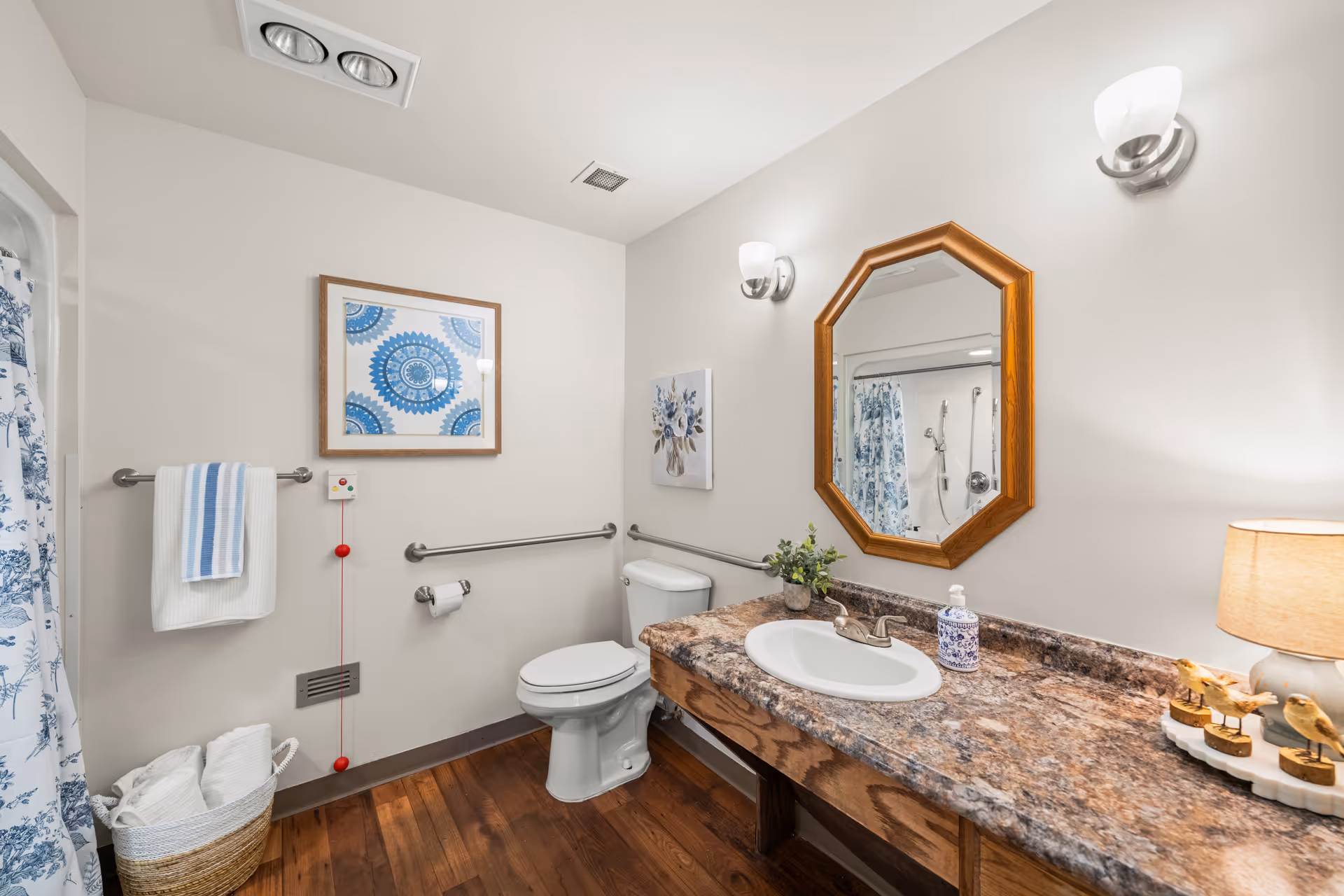 A clean and well-lit bathroom with a white toilet, a wooden vanity with a marble countertop, a white sink, and an octagonal wooden-framed mirror above it. There are two wall-mounted light fixtures, a small table lamp, a basket with towels, and two framed artworks on the walls. The bathroom has wooden flooring and a shower with a blue and white patterned curtain.