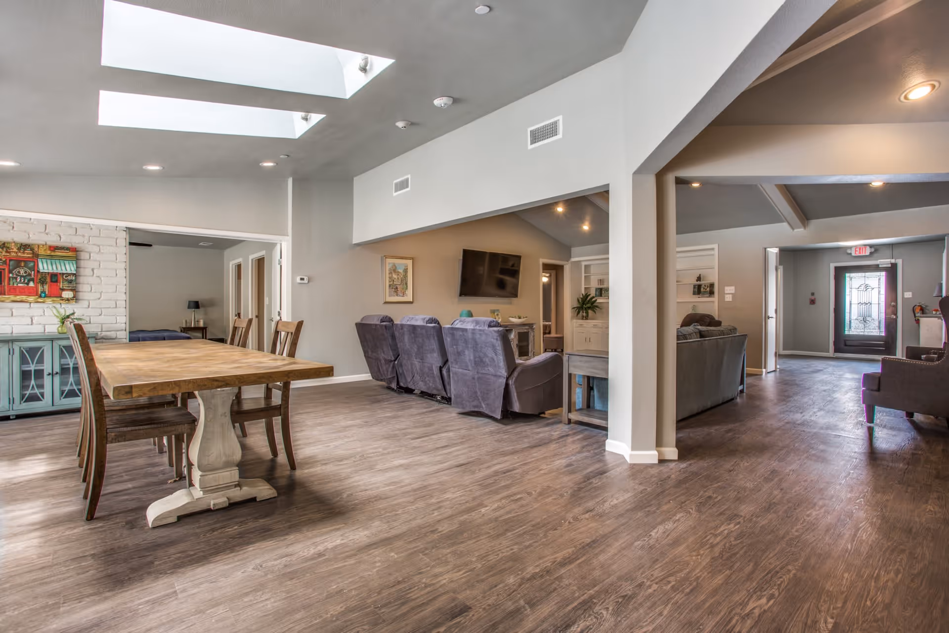 Interior view of a senior living facility showing a spacious room with a wooden dining table and chairs on the left, a seating area with recliners and a wall-mounted TV in the center, and a hallway leading to a glass door exit on the right. The room has wooden flooring, light gray walls, and skylights on the ceiling.