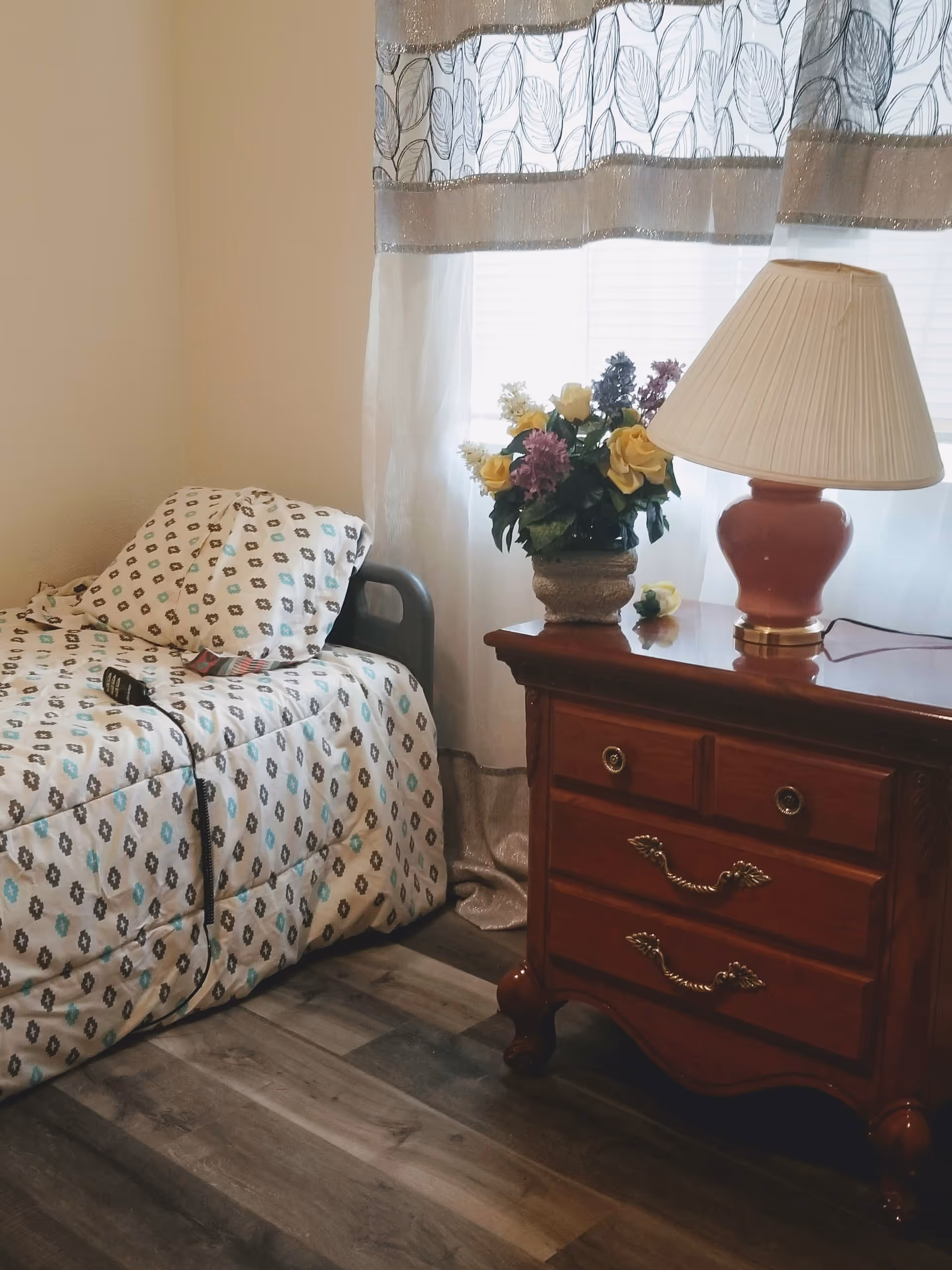 Cozy bedroom with a patterned bed, wooden nightstand, lamp, and flower arrangement.