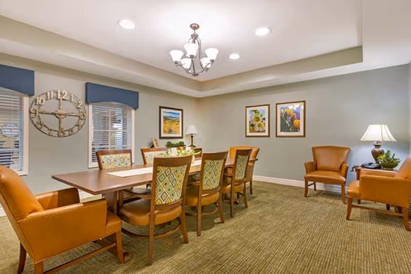 Senior living facility dining room with a long table, chairs, wall art, and warm lighting.