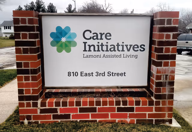 A brick signpost outdoors displaying the logo and name 'Care Initiatives Lamoni Assisted Living' with the address 810 East 3rd Street. The sign has a colorful flower-like logo on the left side and is situated on a grassy area with a sidewalk and parked vehicles visible in the background.