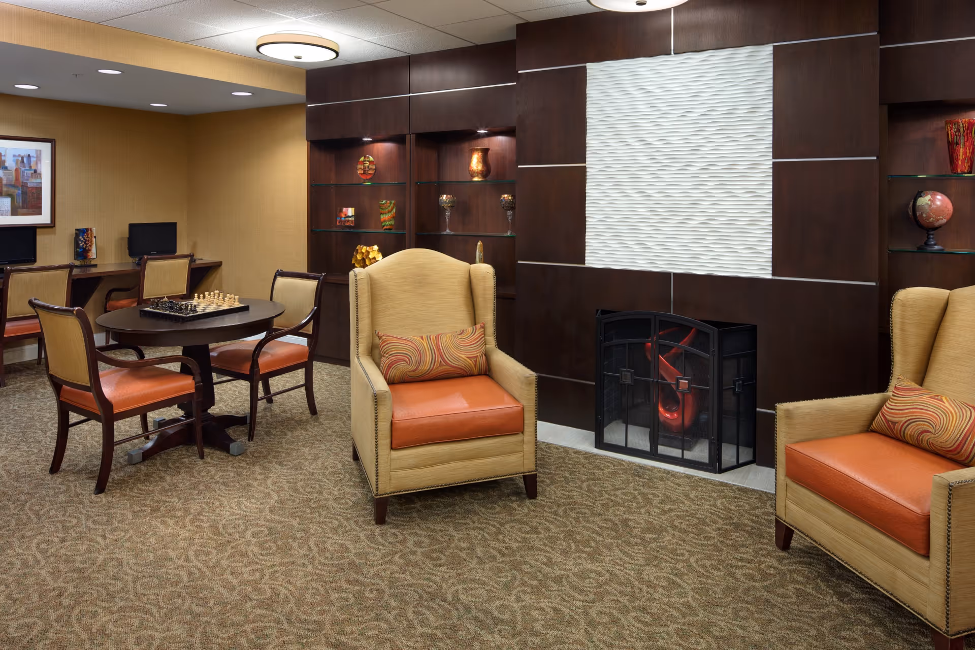 Cozy lounge with chairs, a fireplace, and a card table in a senior living facility.