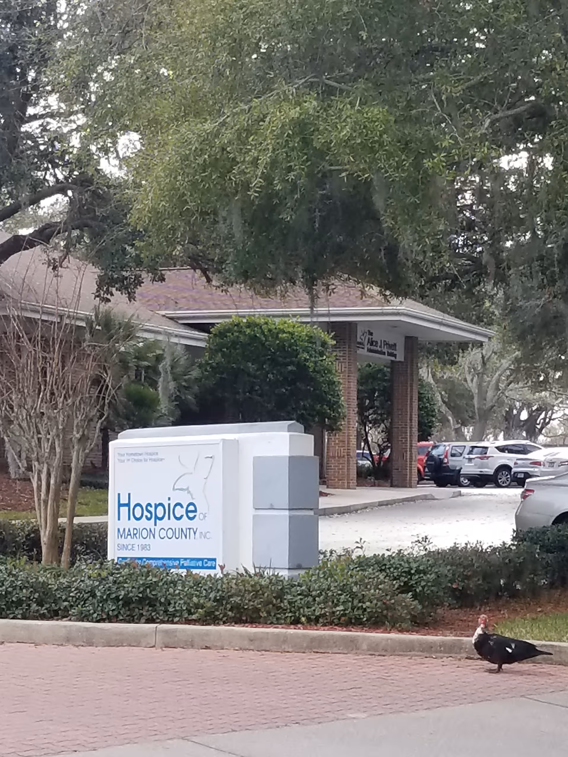 Exterior view of Hospice building with sign, trees, and parked cars.