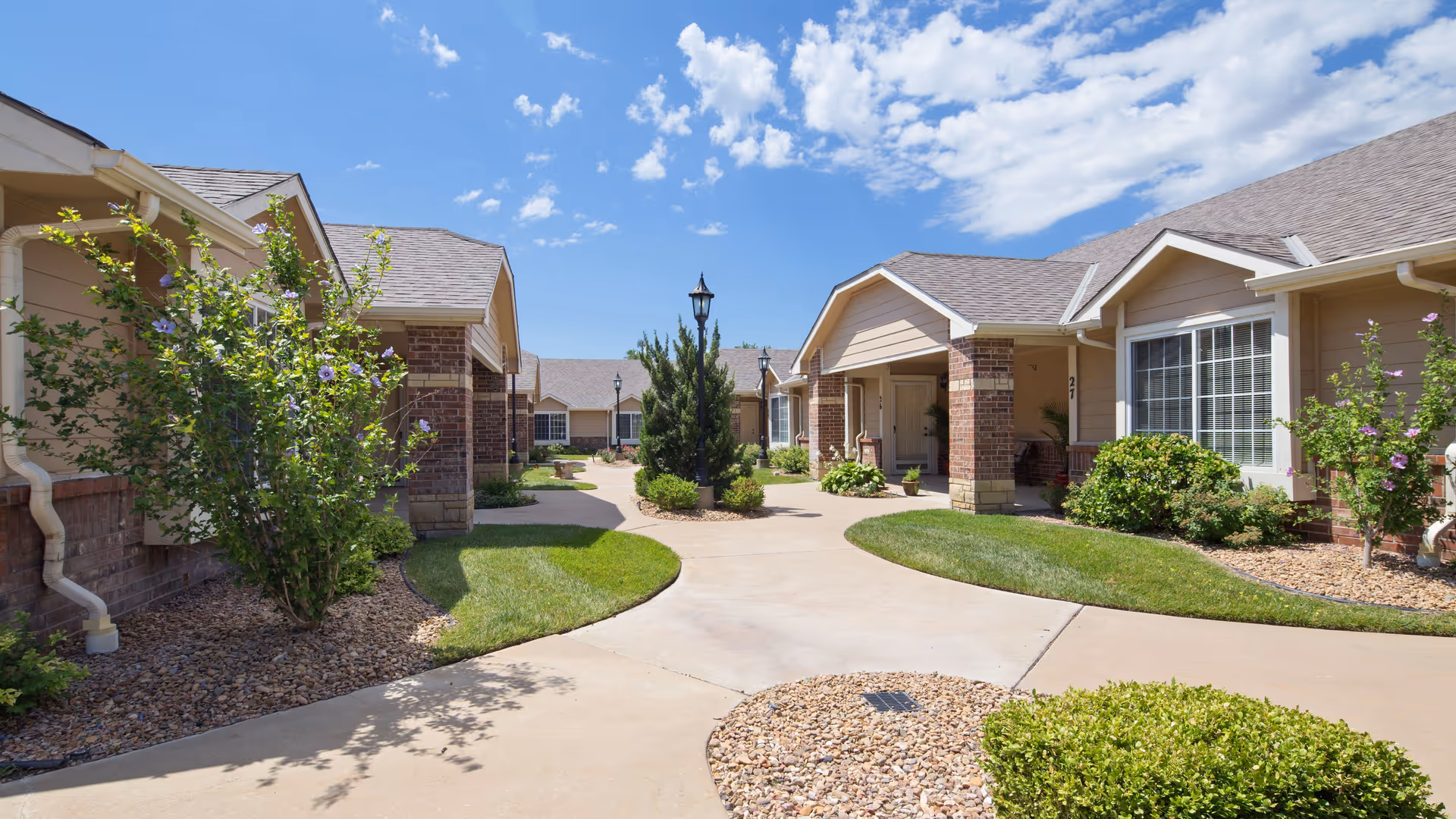 Sunny courtyard of single-story senior living cottages with paved walkways, landscaping, and lamp posts.