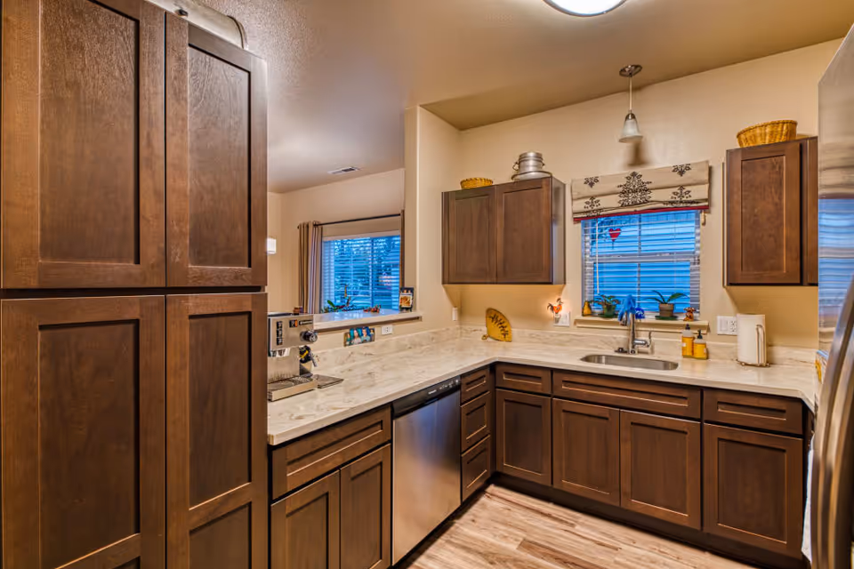 A modern kitchen with dark wooden cabinets, a marble countertop, a stainless steel dishwasher, and a sink under a window with a decorative valance. The kitchen has light-colored walls and wood flooring, with some small plants and kitchen items on the countertop.