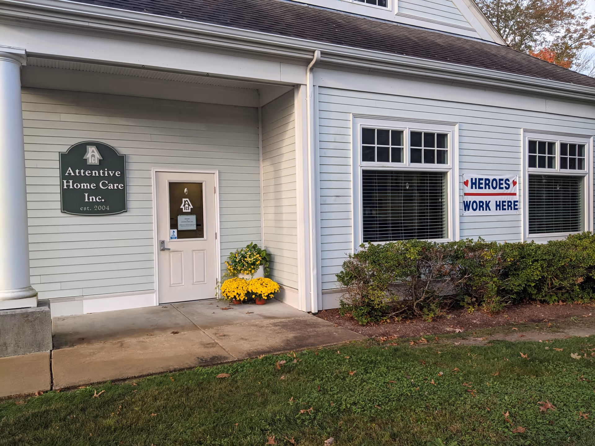 Exterior view of Attentive Home Care building front with signs, door, and yellow potted flowers.