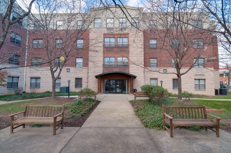 Front entrance of a multi-story brick and stucco senior living building with benches and a walkway.