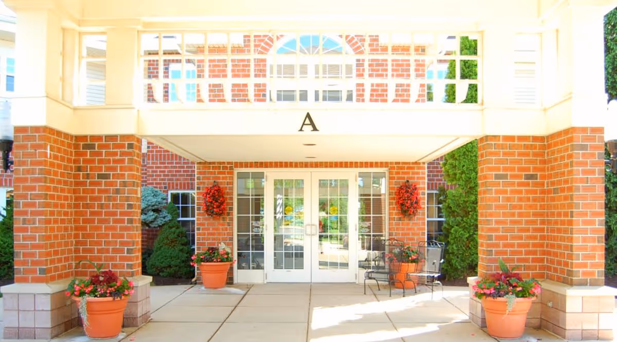 Entrance to a building with double glass doors framed by red brick walls and white trim. There are two large brick columns supporting a covered walkway with a white railing above. Potted plants with flowers and greenery are placed on either side of the entrance, along with two black metal chairs on the right side. The letter 'A' is displayed above the doorway.