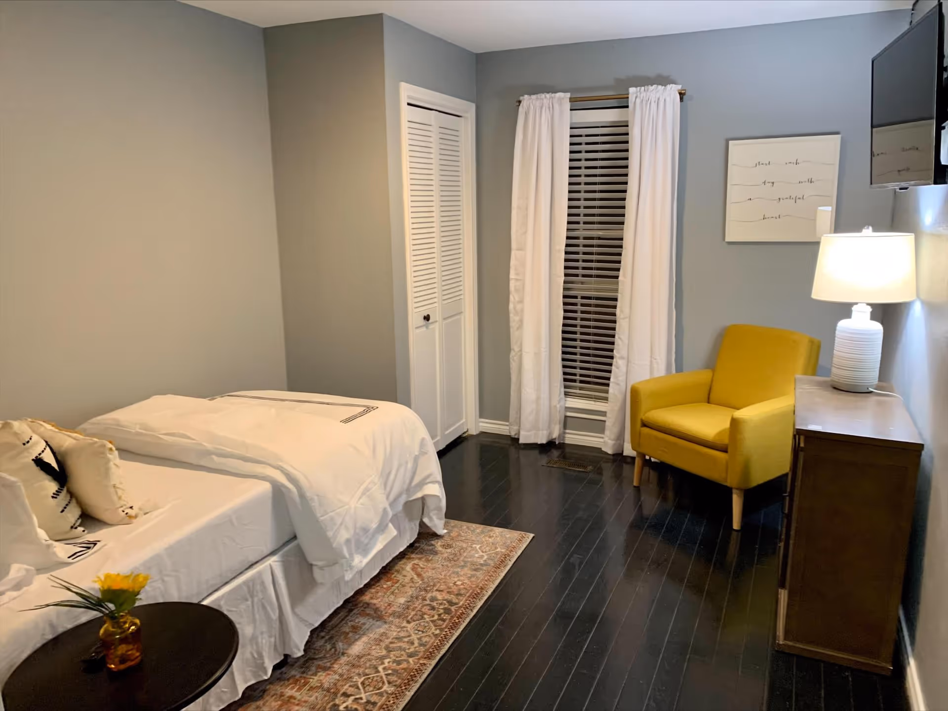 A cozy bedroom with a single bed covered in white bedding and decorative pillows. Next to the bed is a small round table with a small vase holding a yellow flower. The room has dark wooden floors with a patterned rug partially under the bed. A yellow armchair sits near a window with white curtains and blinds. A wooden dresser with a white lamp and a wall-mounted TV are on the right side of the room. A framed piece of art hangs on the wall above the armchair.