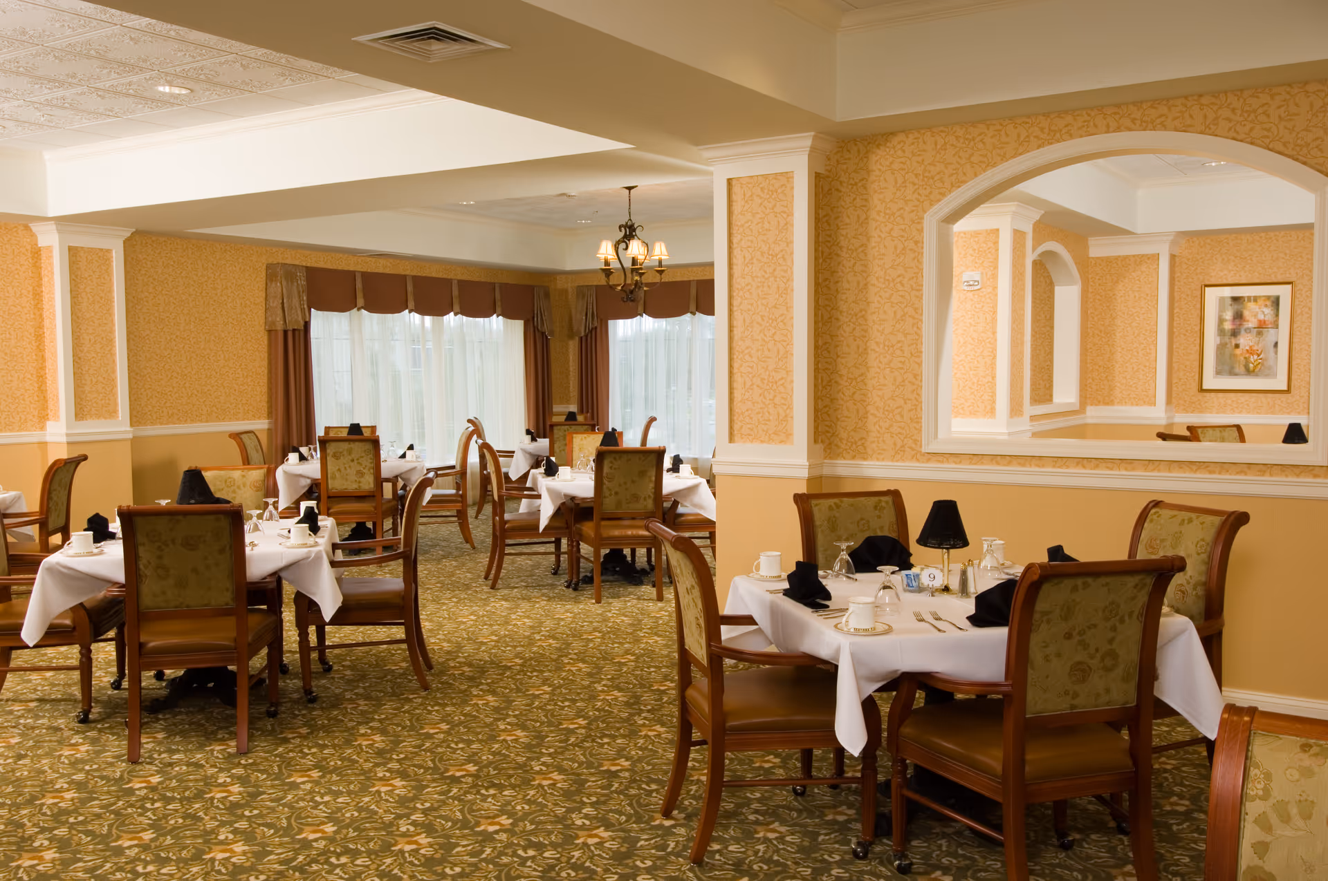 Formal dining room with multiple tables set with white tablecloths, wooden chairs, chandeliers, and patterned carpet.