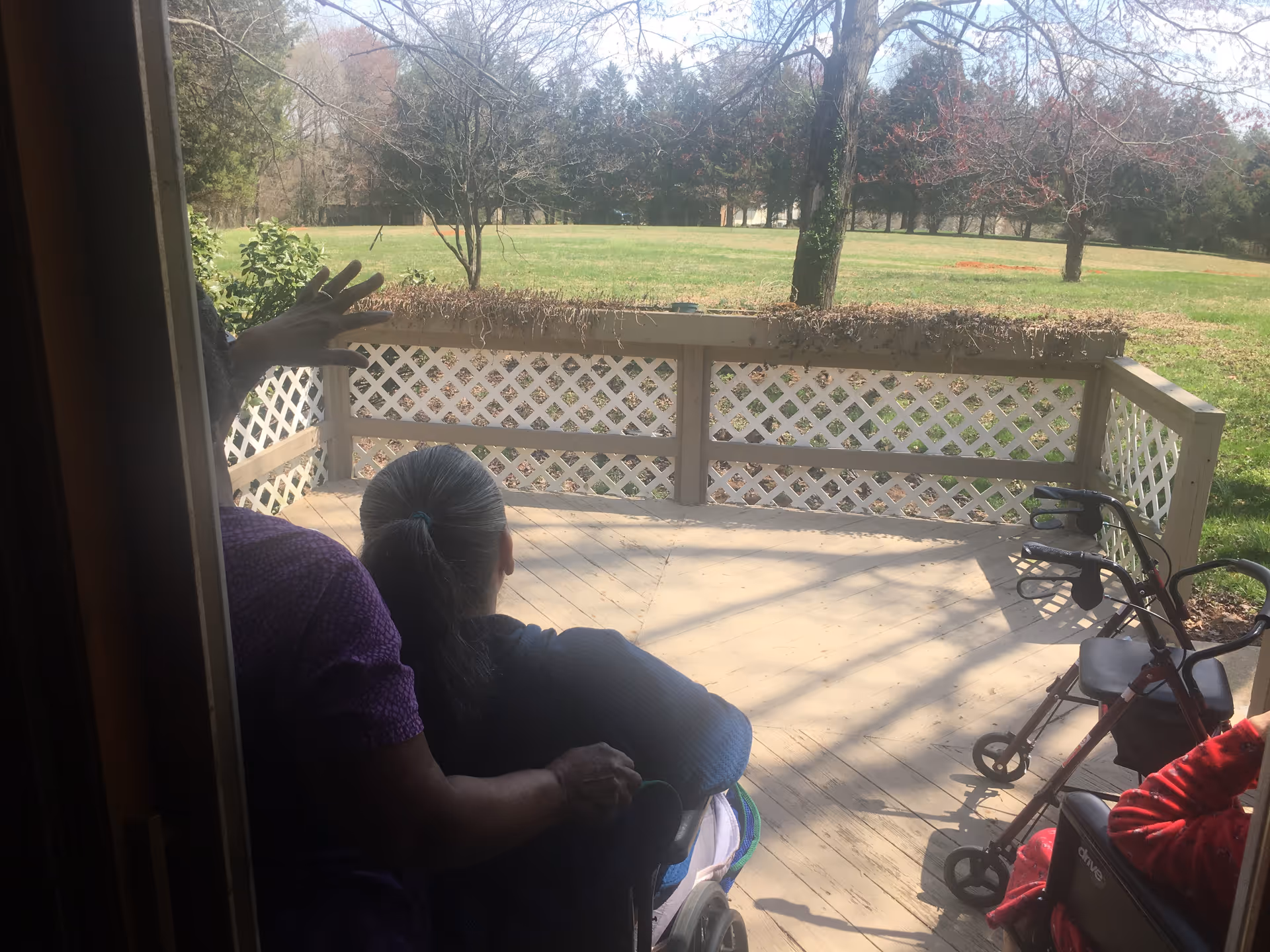 View from inside a building looking out onto a wooden deck with white lattice railing. Two elderly individuals are seated in wheelchairs on the deck, one person is standing behind and gently touching one of the seated individuals. The deck overlooks a grassy area with trees in the background under a clear sky.