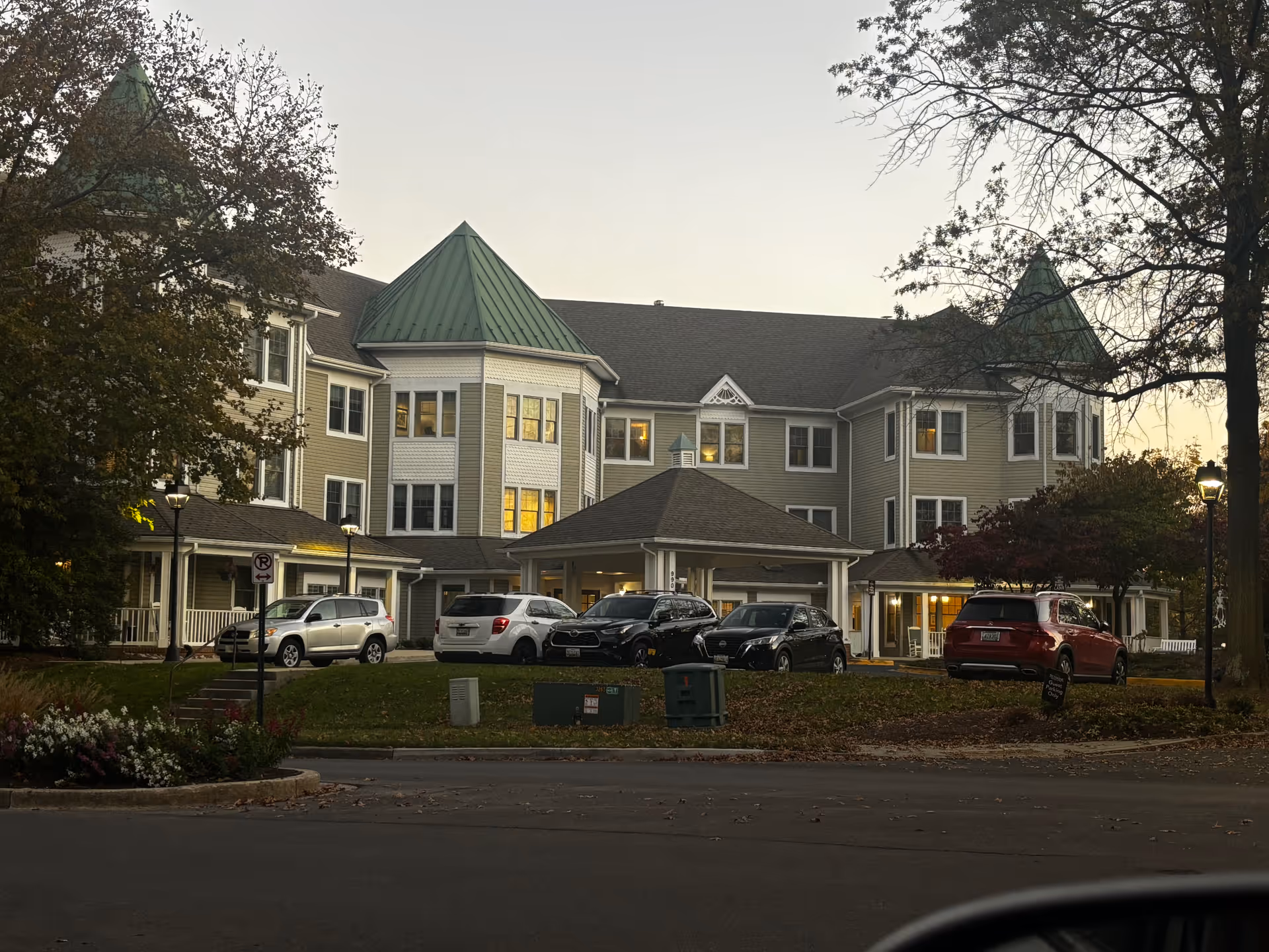 Front exterior of a three-story residential building with green turret roofs, lit windows, and parked cars at dusk.