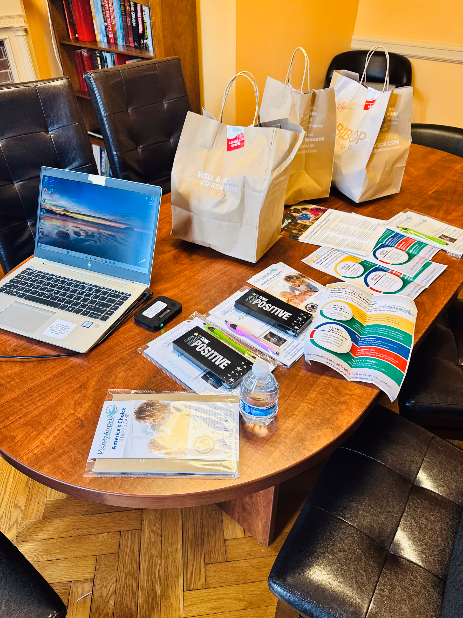 A wooden conference table with laptop, brochures, bags, water bottle, and chairs in a bright room.