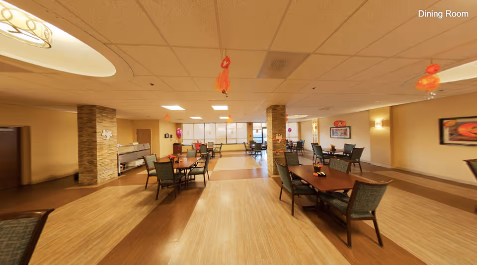 A spacious dining room with multiple wooden tables and chairs arranged neatly. The room has a warm color scheme with beige walls and a tiled ceiling. There are decorative Halloween-themed hanging ornaments and wall decorations. Large windows at the far end allow natural light to brighten the space.