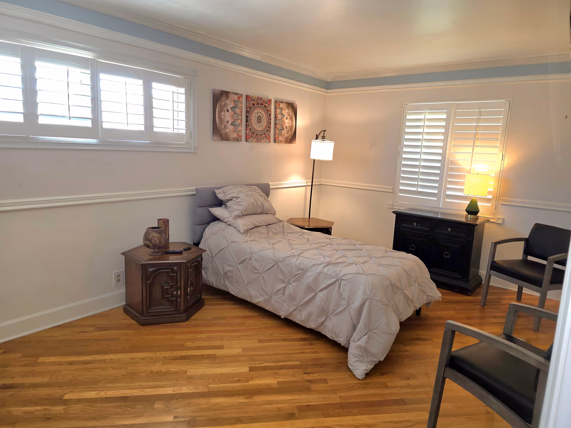A simple bedroom with a single bed covered in a light gray comforter and two pillows. The room has wooden floors, white walls with a chair rail, and two windows with white plantation shutters. There is a wooden nightstand with a decorative item and a remote control, a floor lamp beside the bed, a black cabinet with a green lamp on top, and two black chairs with wooden armrests.