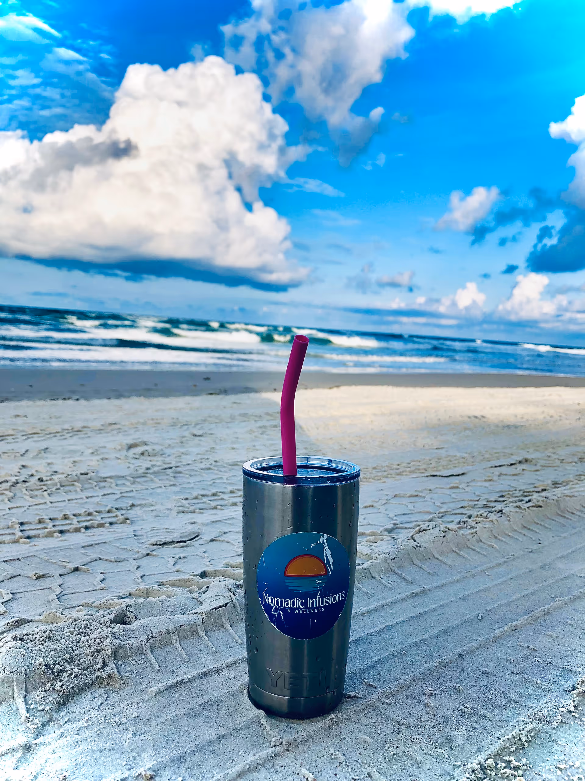 A stainless tumbler with purple straw and Nomadic Infusions logo sits on a sunny beach.