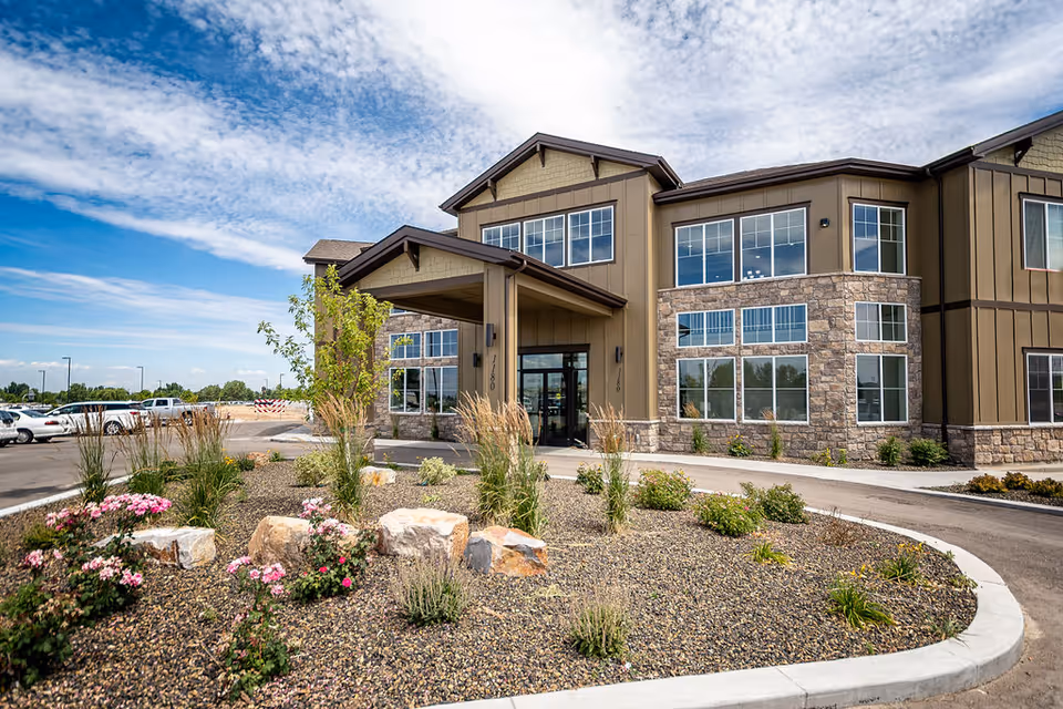Exterior view of a two-story senior living facility building with large windows and a covered entrance. The landscaped area in front features rocks, flowering plants, and small shrubs under a partly cloudy blue sky.