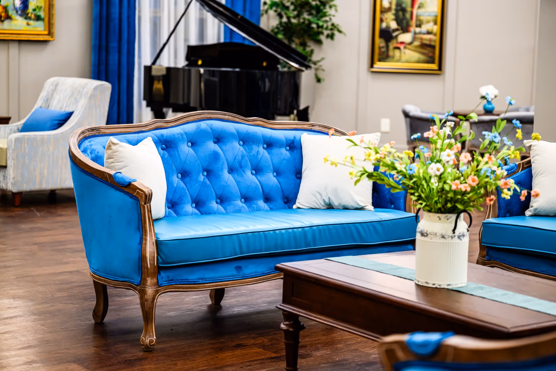 A bright communal lounge with a blue tufted sofa, coffee table holding a vase of flowers, and a grand piano in the background.