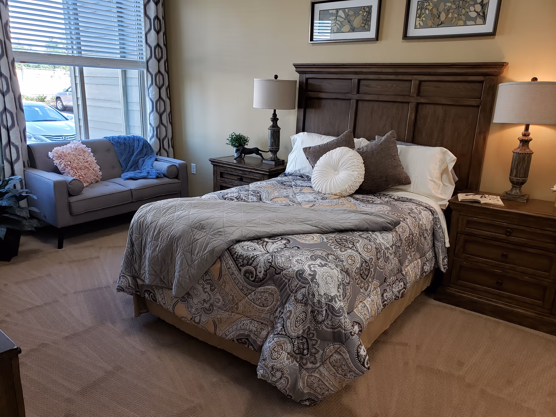 A furnished bedroom with a bed, wooden headboard, nightstands with lamps, and a small sofa by the window.