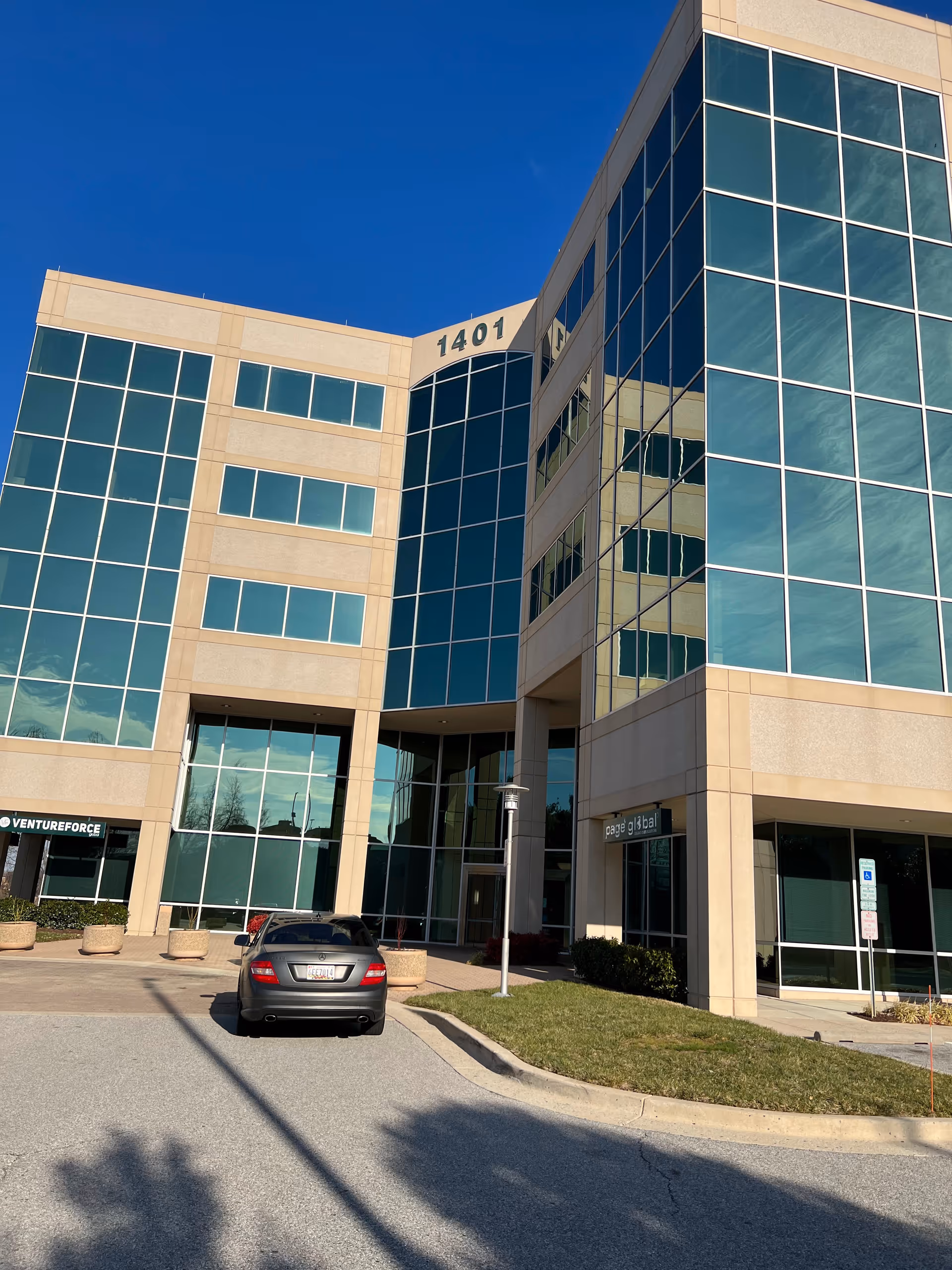 Modern glass-front office building exterior with beige trim, a parked car, and blue sky.