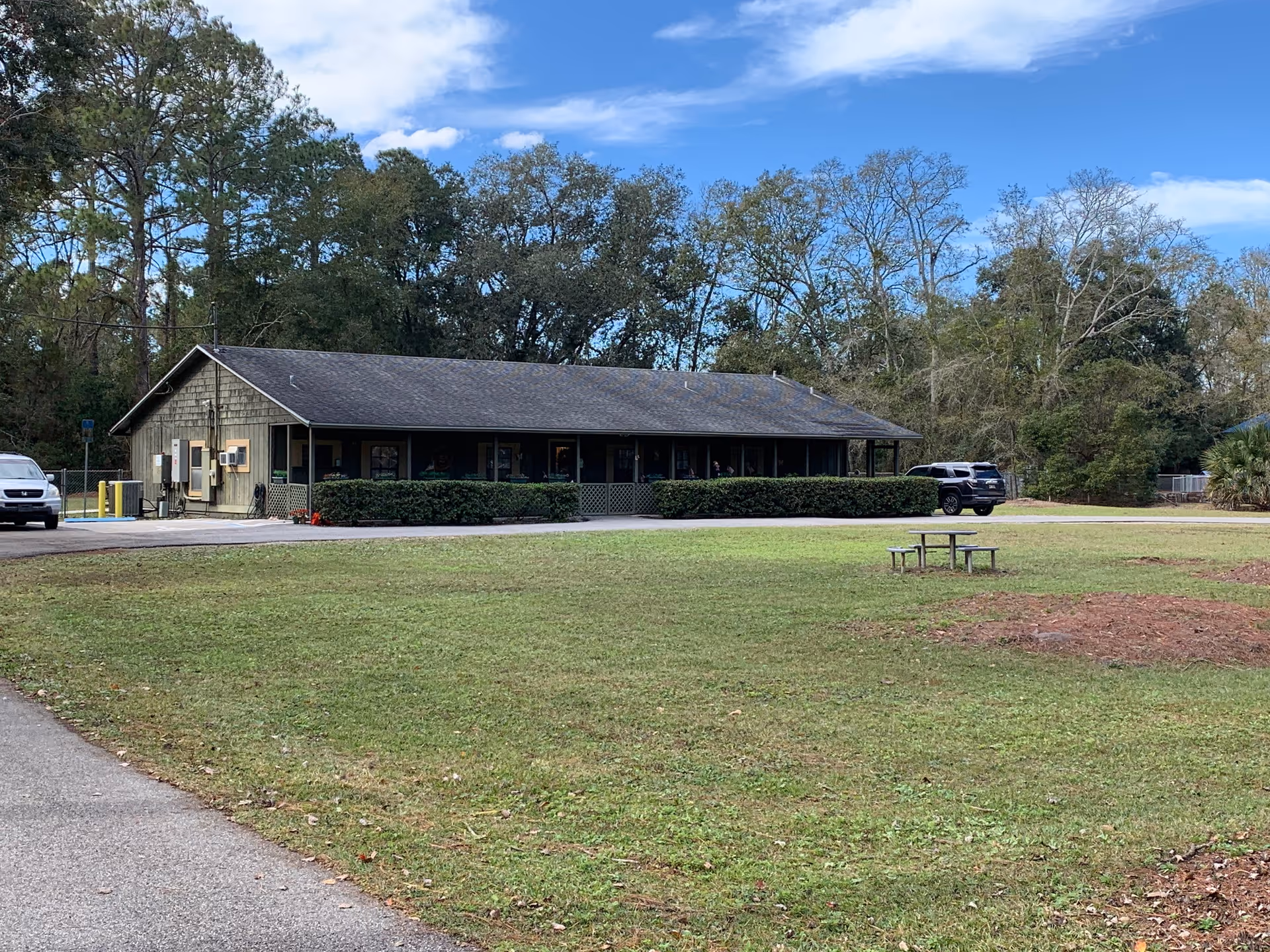 Single-story building with a dark roof and greenish exterior walls surrounded by trees and a grassy lawn with a picnic table. Two vehicles are parked near the building under a partly cloudy blue sky.