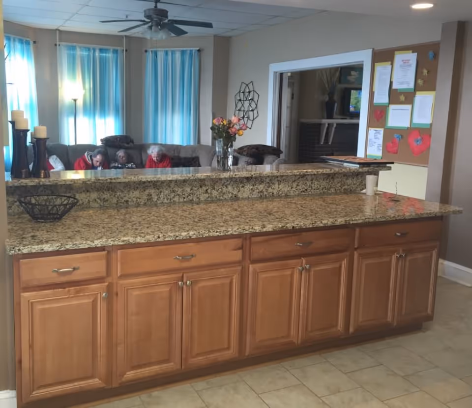 Granite countertop with wooden cabinets dominates foreground; residents sit on sofas in a living area behind.
