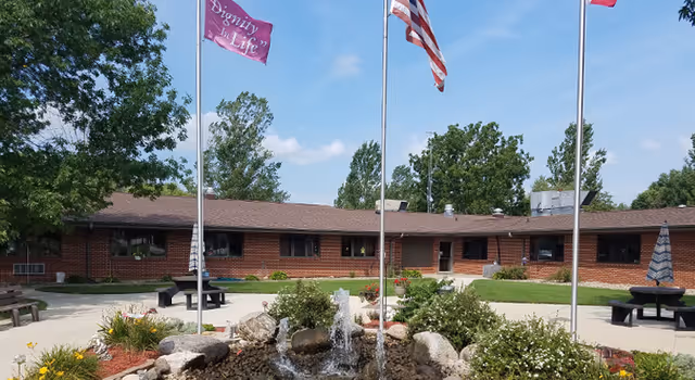 Outdoor courtyard area of a senior living facility with a central water fountain surrounded by rocks and plants. There are three flagpoles with flags, including an American flag and a pink flag that reads 'Dignity in Life'. The courtyard is paved with benches and tables, and a single-story brick building with windows and a brown roof surrounds the area. Trees and a blue sky with some clouds are visible in the background.