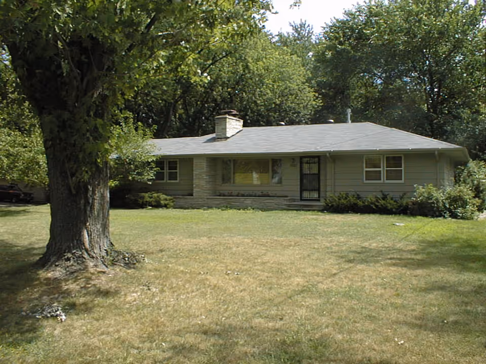 Single-story gray building with a metal door, surrounded by trees and a grassy yard.