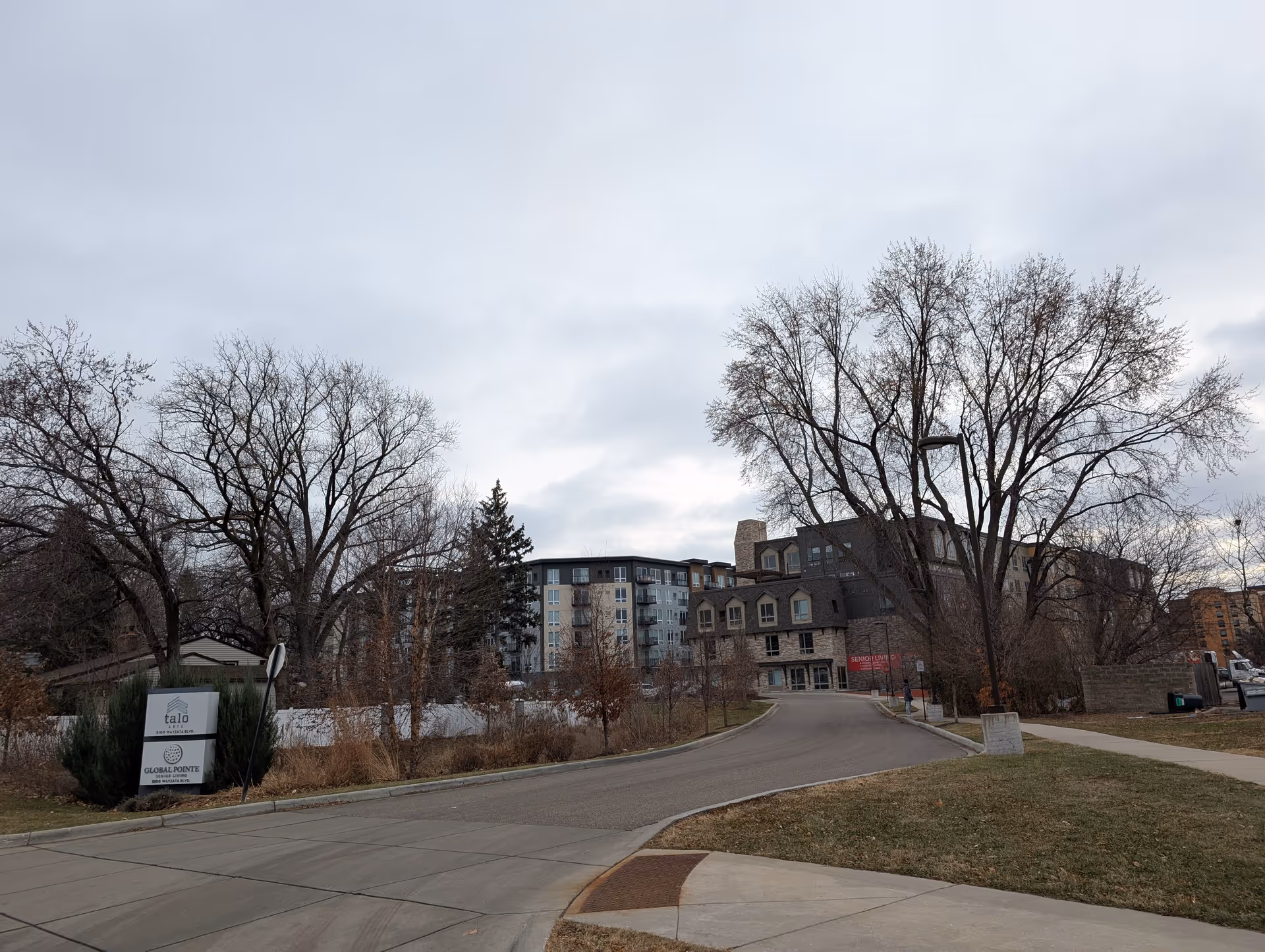 Exterior view of the Global Pointe Senior Living building and driveway framed by leafless trees and lawn under a cloudy sky.