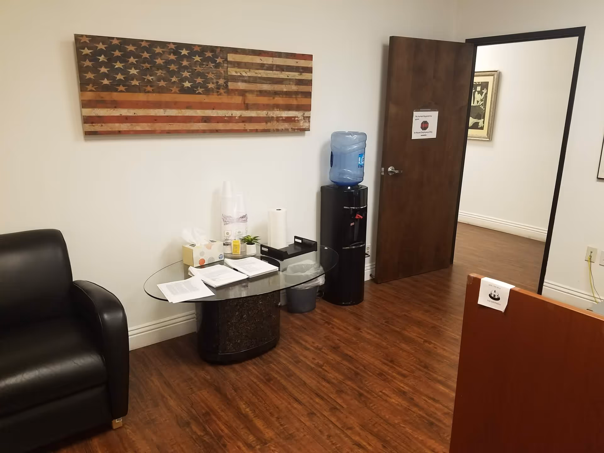 Interior waiting area featuring a glass round table, chair, water cooler, and flag artwork.