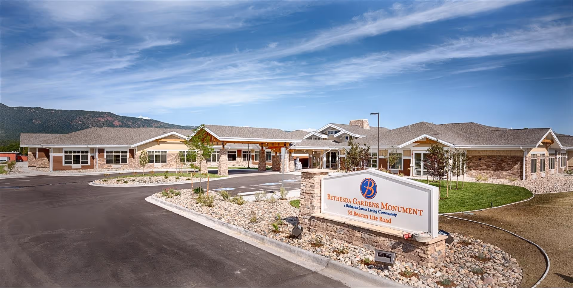 Exterior view of Bethesda Gardens Monument senior living community building with a clear blue sky and mountains in the background. The building has a stone and siding facade with a covered entrance and a landscaped area with rocks and small trees. A sign in front reads 'Bethesda Gardens Monument, A Bethesda Senior Living Community, 55 Beacon Lite Road.'