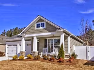 Single-story house with a front porch featuring two white rocking chairs, a double garage, and a fenced yard under a clear blue sky.