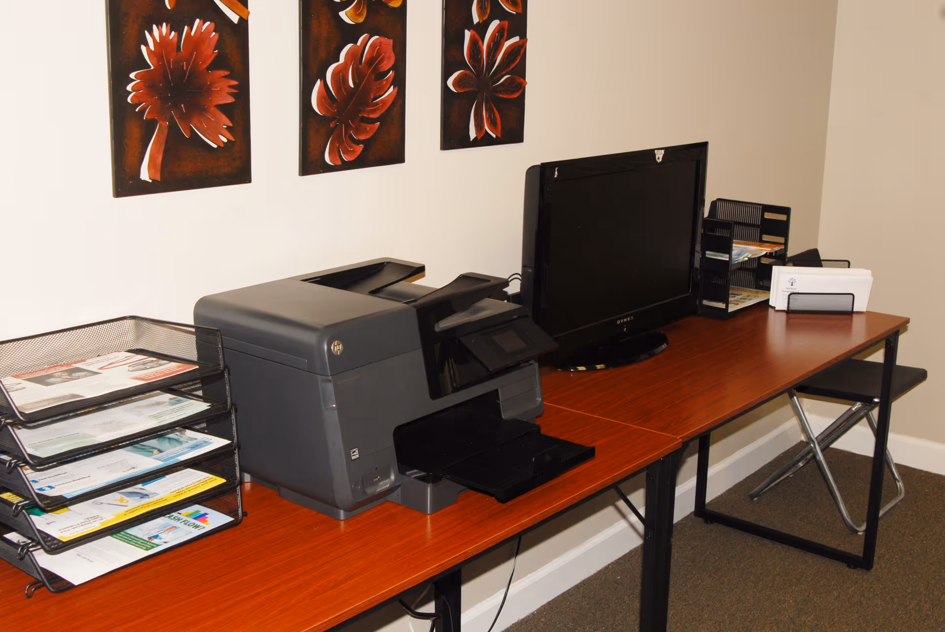 A wooden desk with printer, monitor, and filing trays in an office corner.