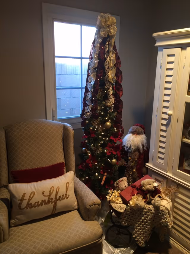 Cozy living room corner with an upholstered armchair bearing a 'thankful' pillow beside a tall decorated Christmas tree, Santa figure, and cabinet.