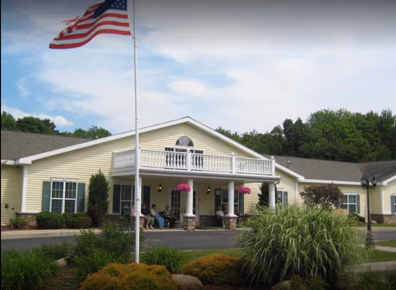 Senior living facility exterior with flagpole, porch, and residents seated outside.