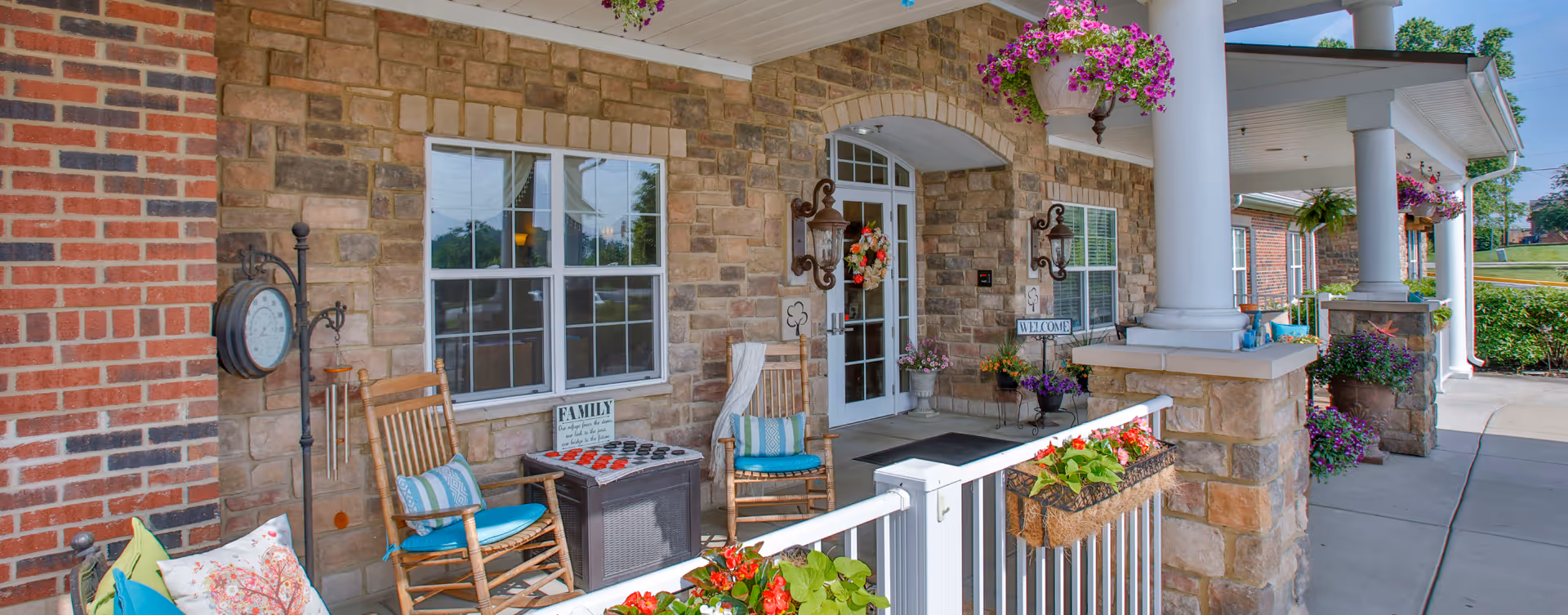 Front porch area of Bickford of Greenwood featuring stone and brick exterior walls, white columns, hanging flower baskets, rocking chairs with cushions, a checkers game on a small table, and a welcoming entrance door with a floral wreath.