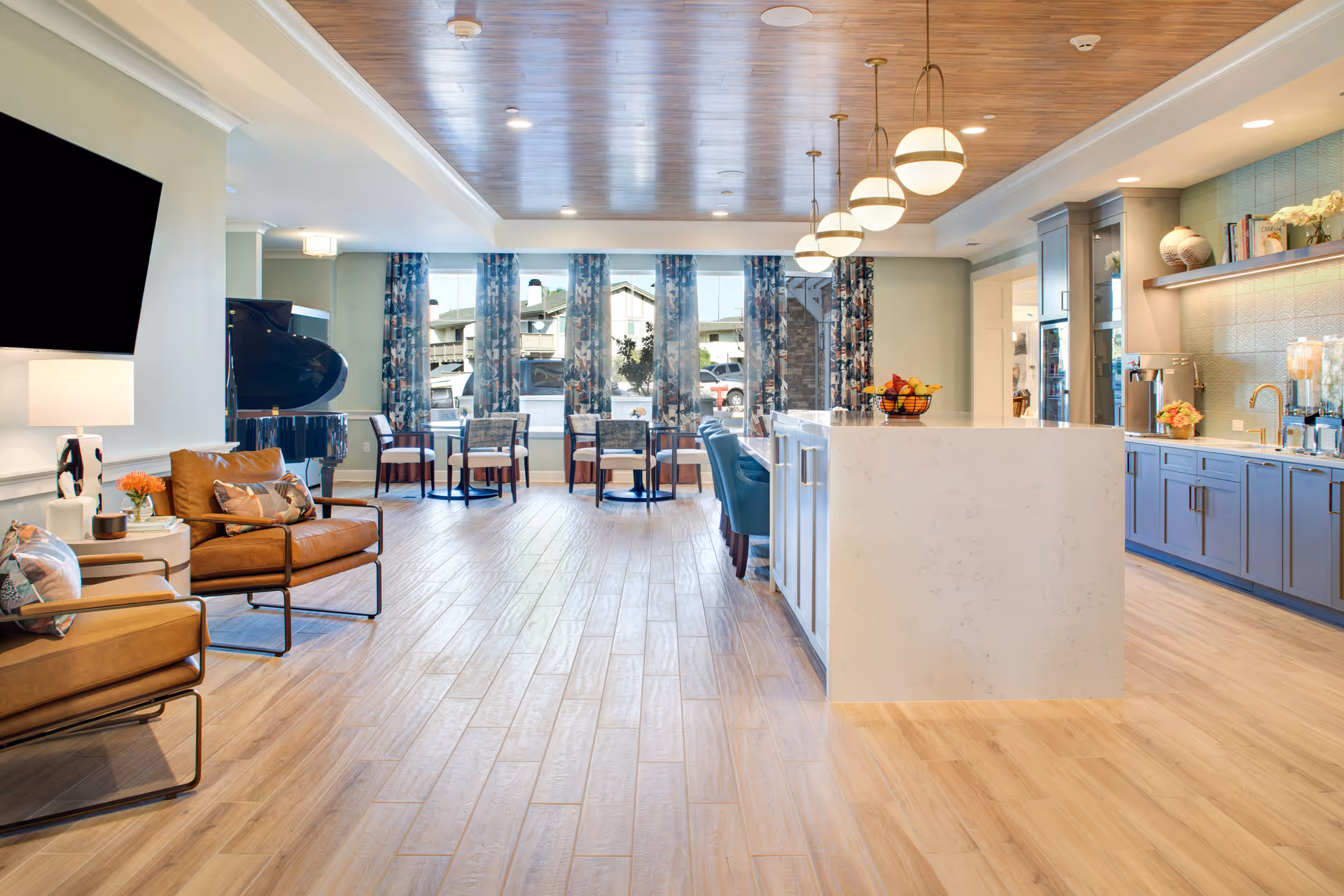 Bright lounge with seating, a kitchen island, and large windows in a senior living facility.