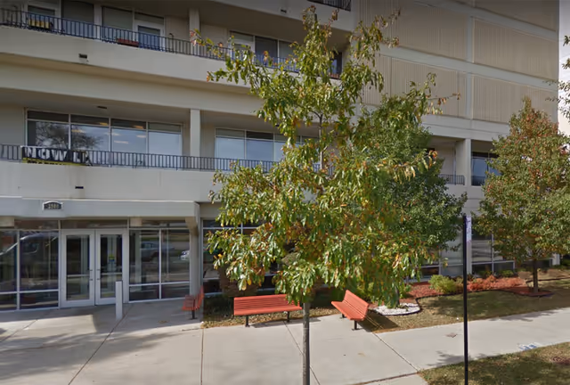 Exterior of Pioneer Gardens senior living facility with glass entrance, trees, and red benches along walkway.