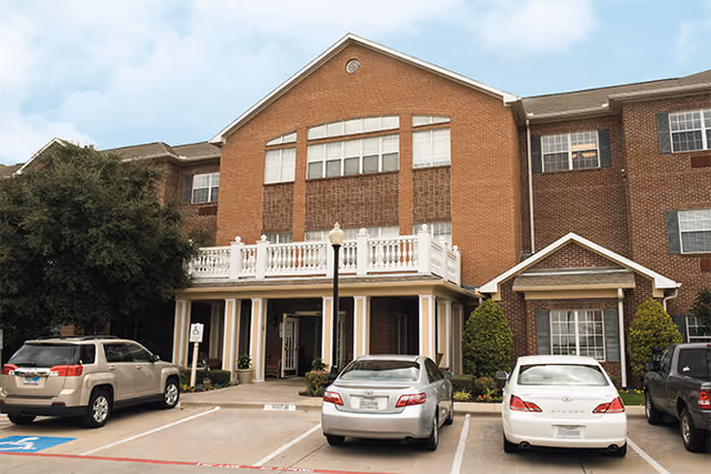 Brick senior living facility with a covered entrance, balcony railings, and parked cars in front.